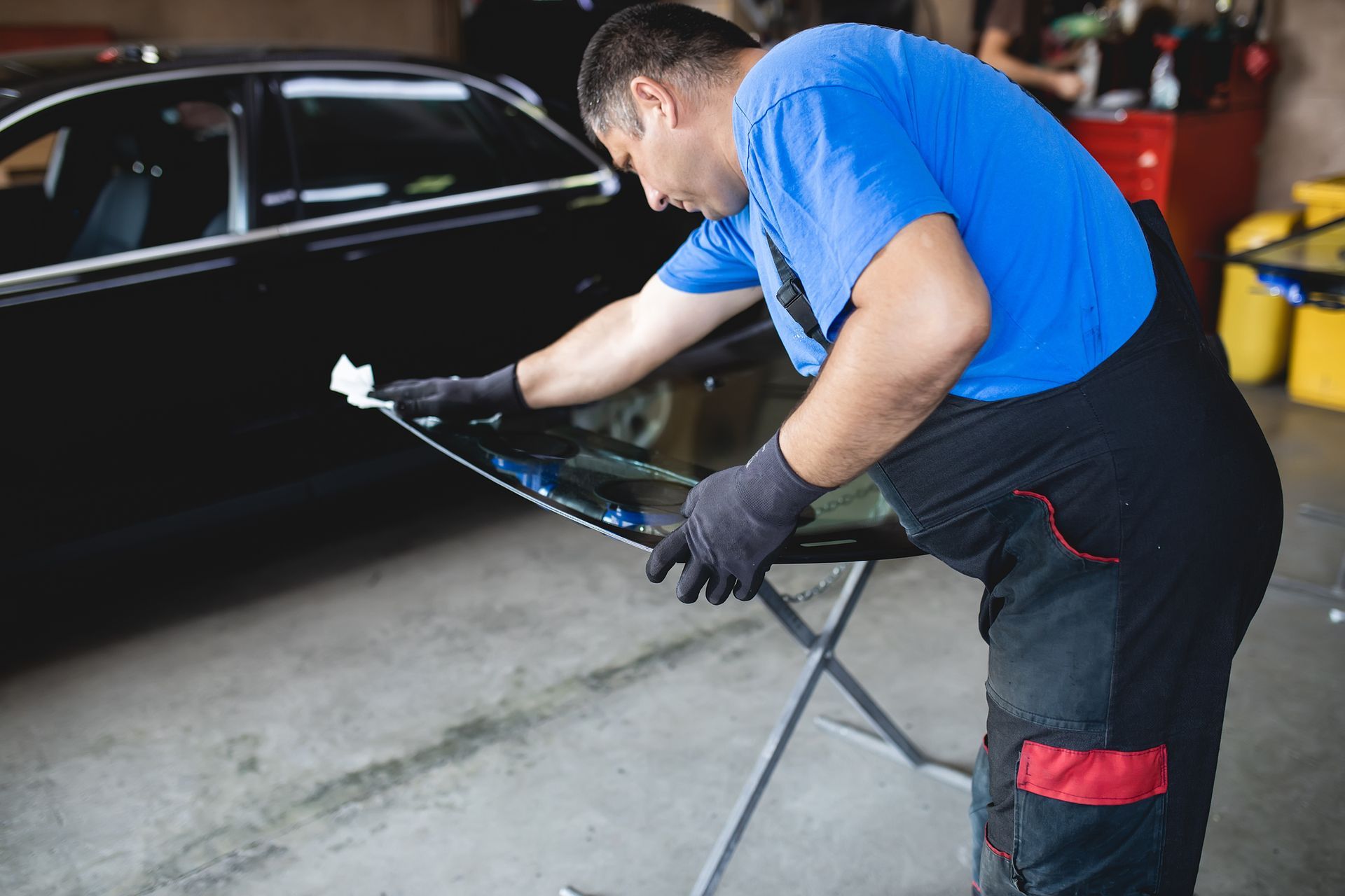 Mechanic cleaning a car windshield in a garage, wearing a blue shirt and black overalls.