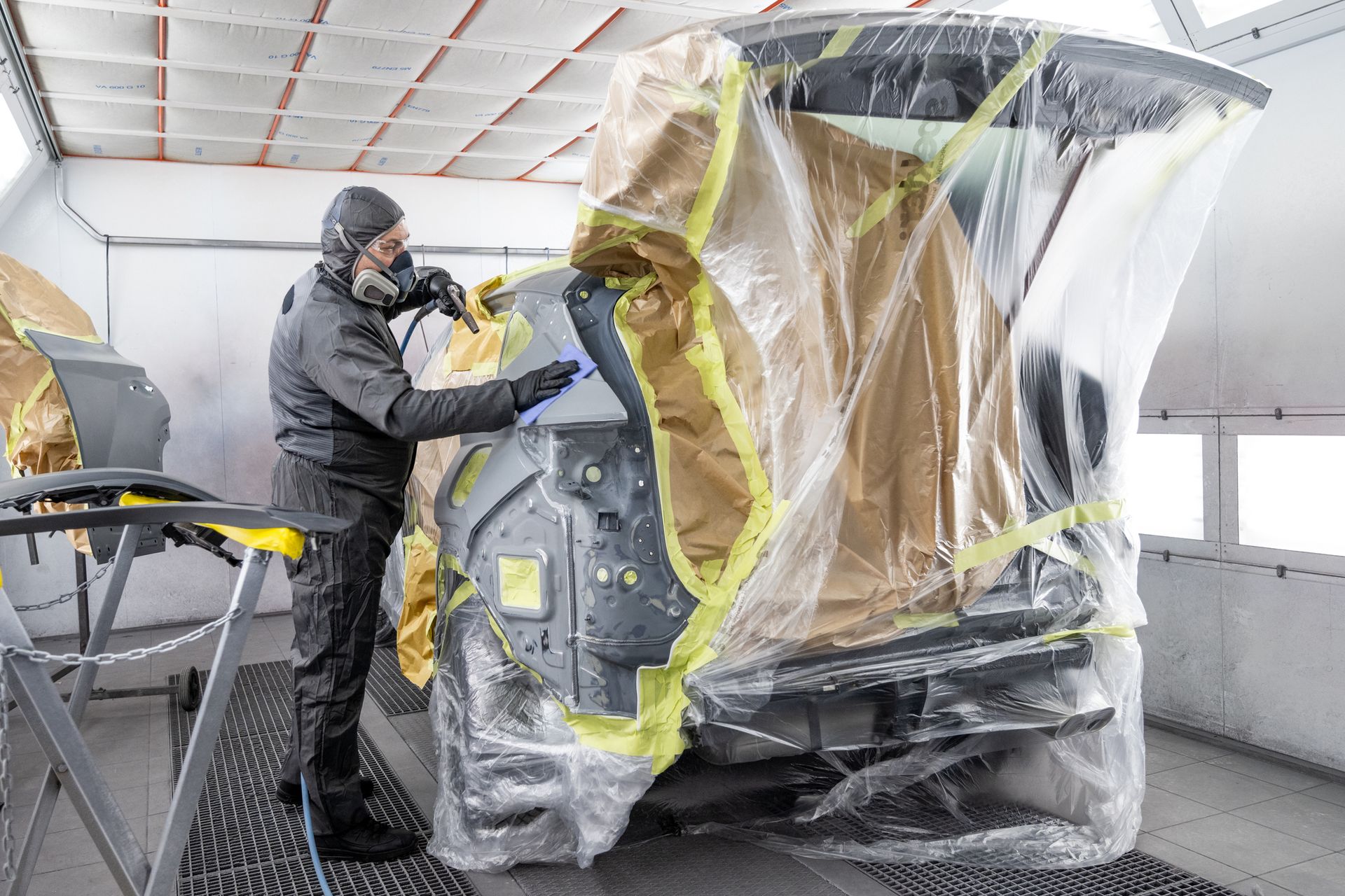 Auto body worker in a paint booth spraying a car panel, wearing protective gear.