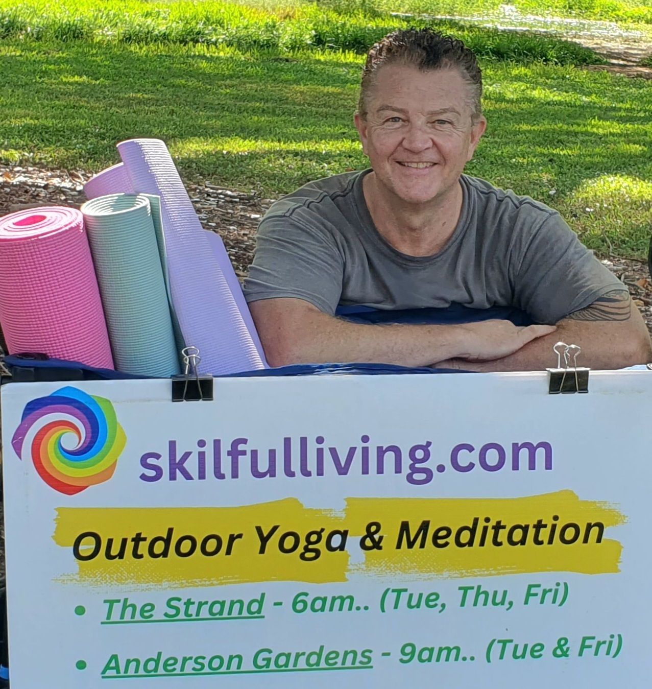 Man smiling behind sign for skilfulliving.com outdoor yoga and meditation classes in a park — Skilful Living in North Ward, QLD