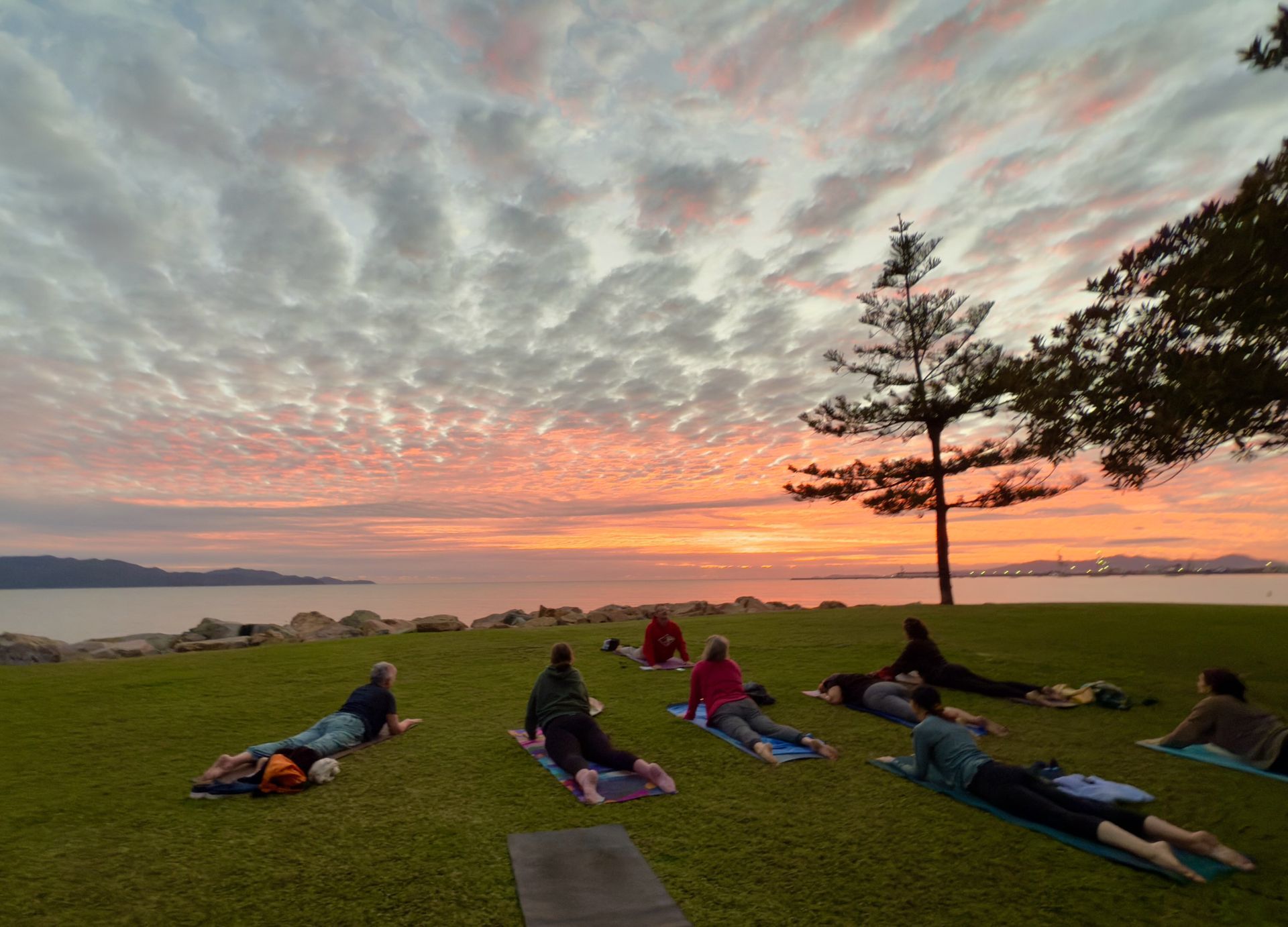 People lying on mats on a grassy hill at sunset overlooking the water and clouds