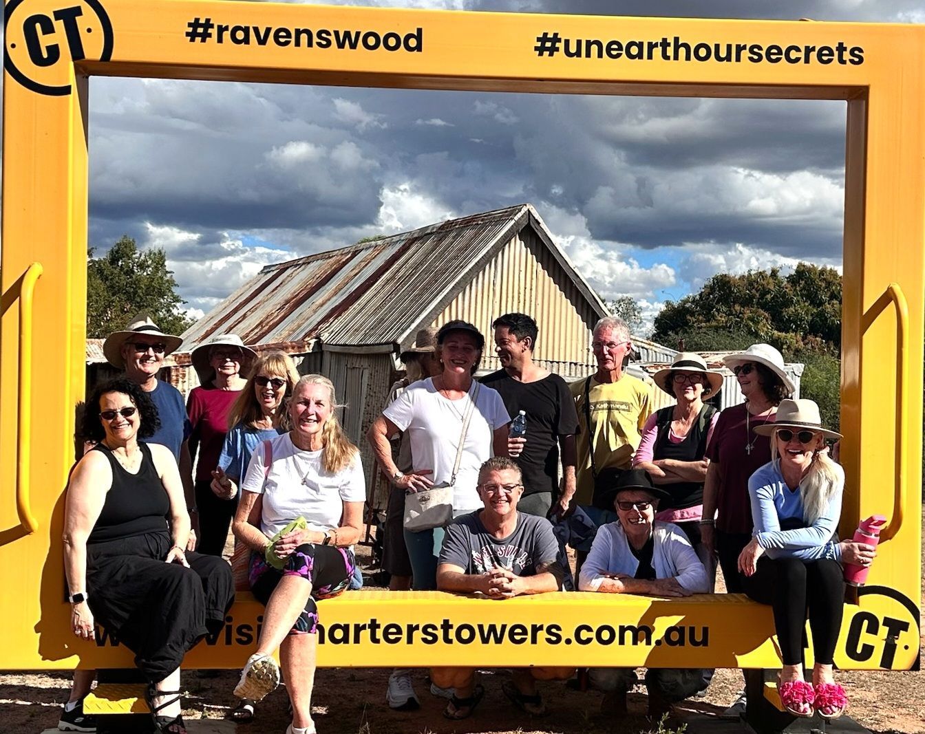 Group posing in front of a rustic house with cloudy sky, framed by a yellow #ravenwood #unearthoursecrets border.