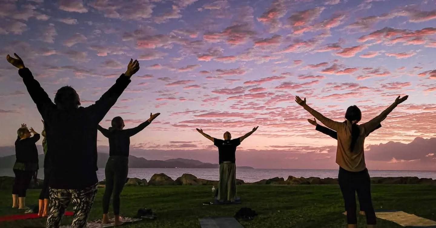 People standing on a grassy hill with arms raised at sunset under a pink cloudy sky