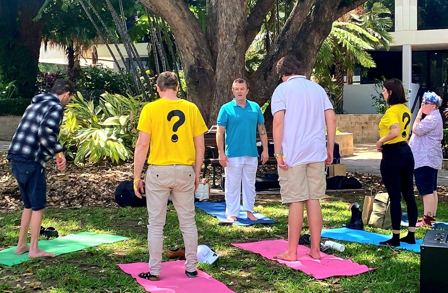 People standing on colourful mats outdoors in a garden, facing a yoga instructor.