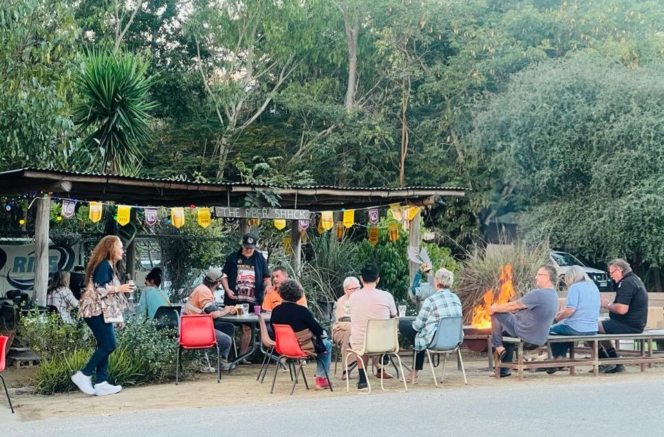 People seated outdoors around a fire pit at a rustic evening gathering under string lights.