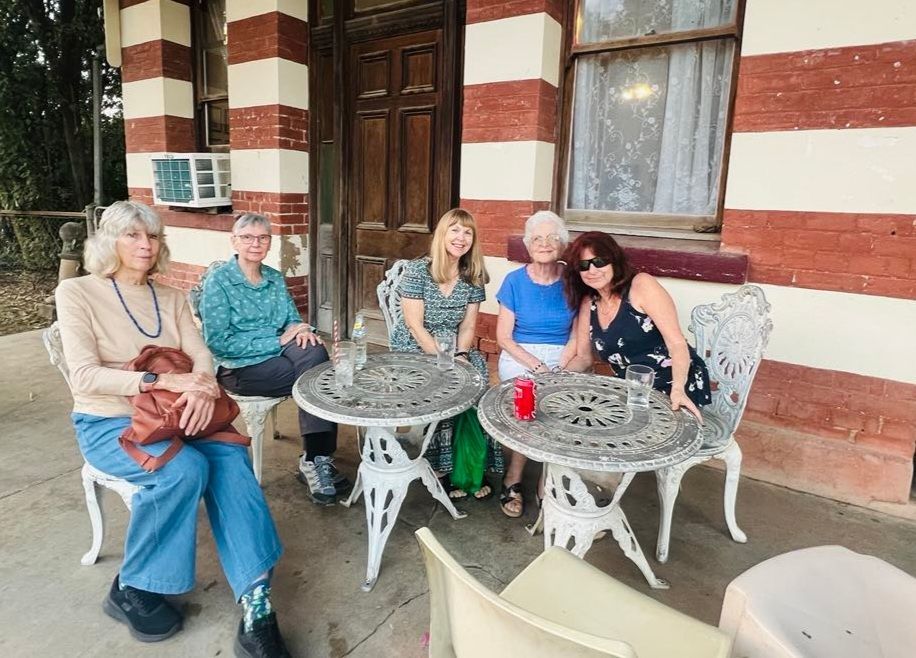 Five people seated at a round outdoor table on a porch with red-and-white striped walls and wooden doors