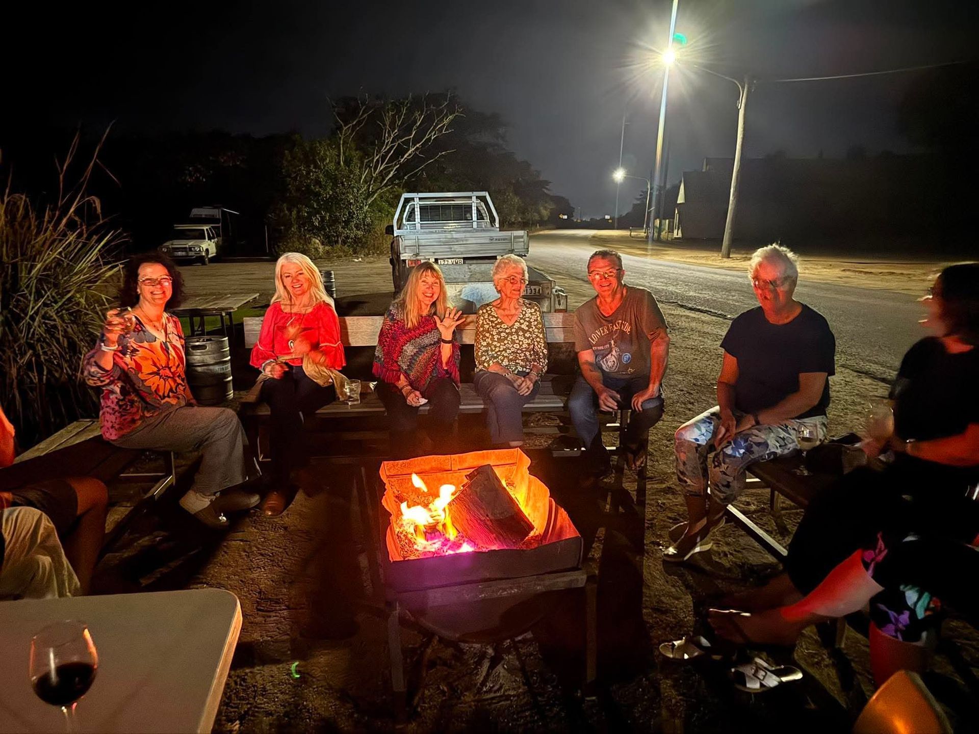 People sitting around a campfire at night under bright lights, with a parked pickup truck nearby.