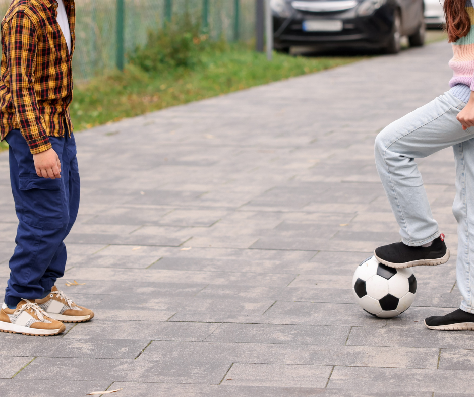 Two people stand on a paved walkway, one resting a foot on a soccer ball.