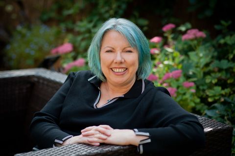 Smiling woman with blue hair sitting outdoors in a wicker chair, with pink flowers in the background