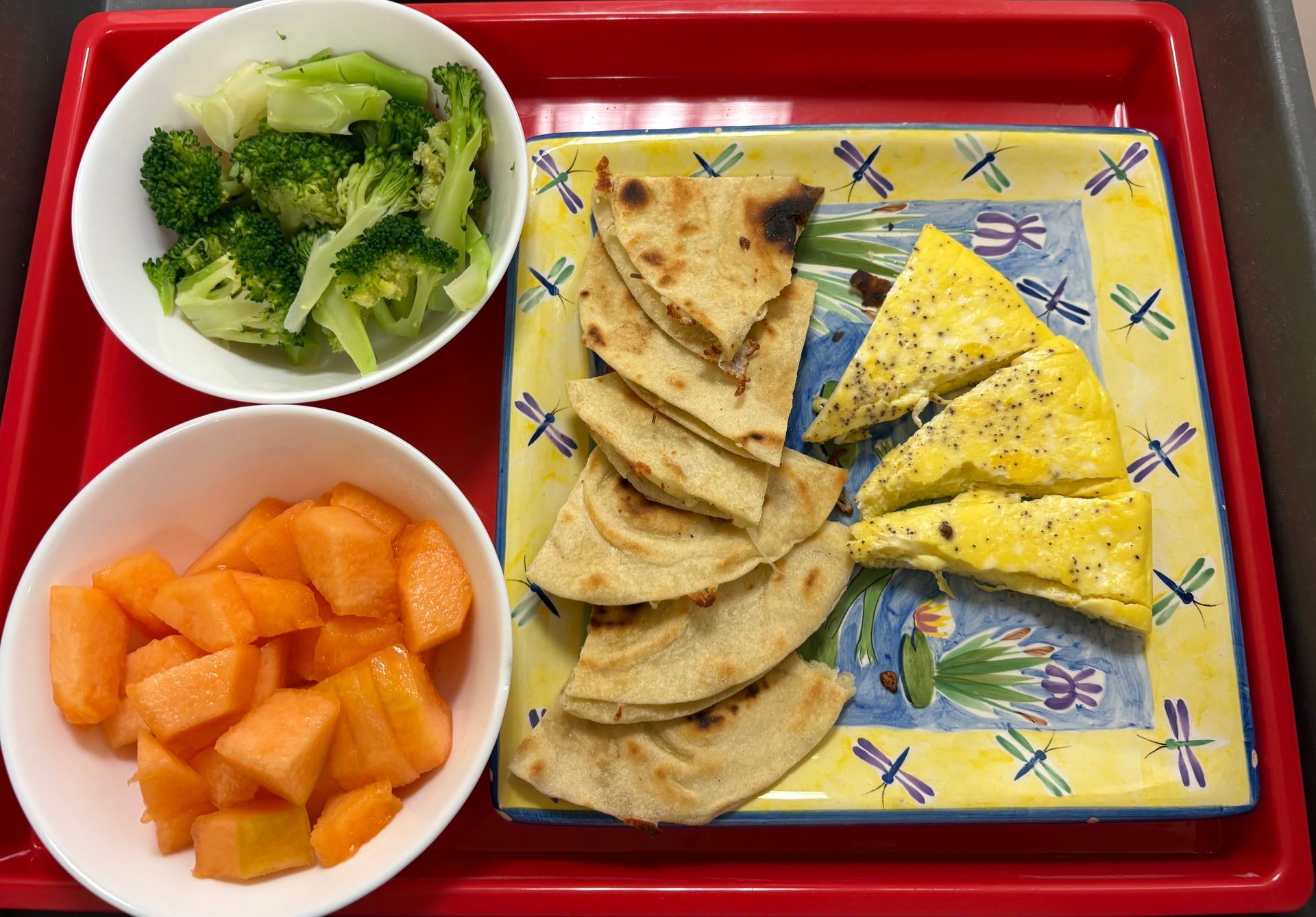 A plate of food with a bowl of broccoli and a bowl of cantaloupe on a red tray.