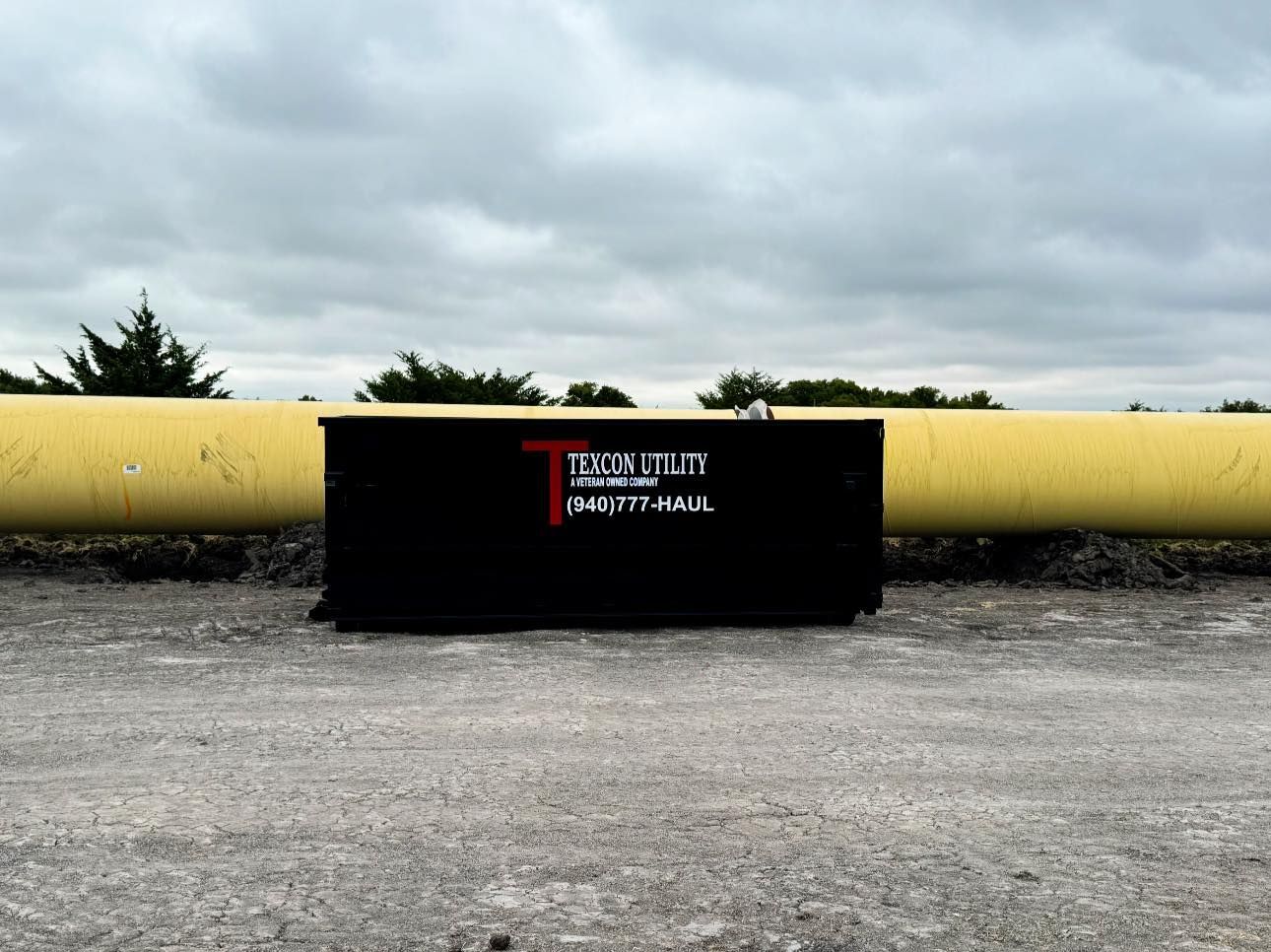 Black dumpster with red logo against a yellow pipe and cloudy sky.