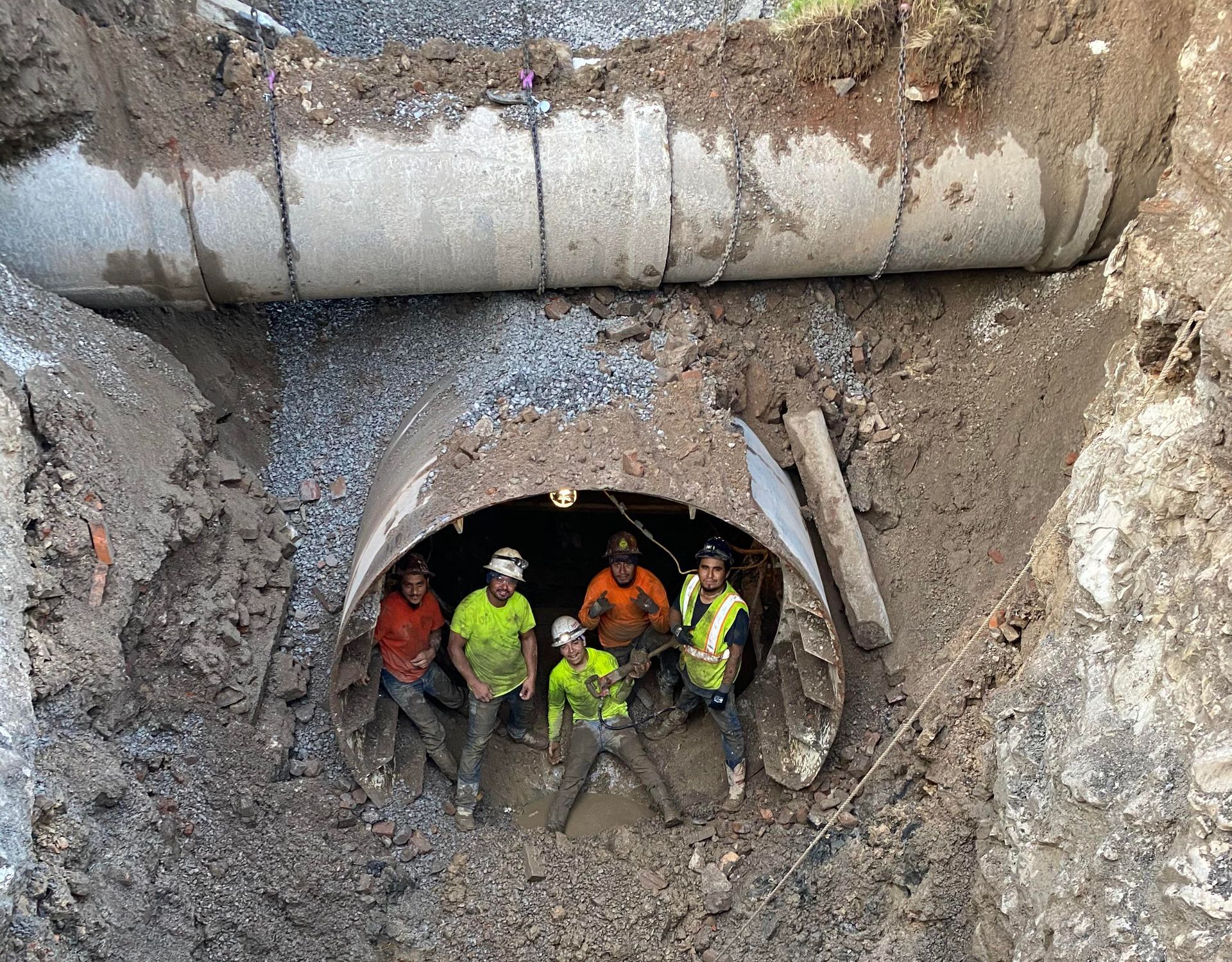 Workers in neon safety gear emerge from a muddy underground tunnel beneath a large pipe