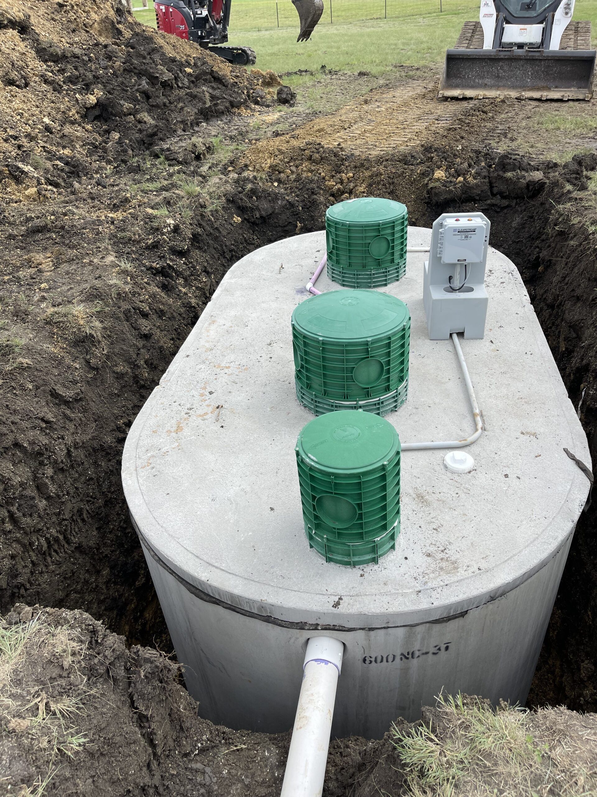 Concrete septic tank in a trench, with green lids and electrical box. Excavator in the background.