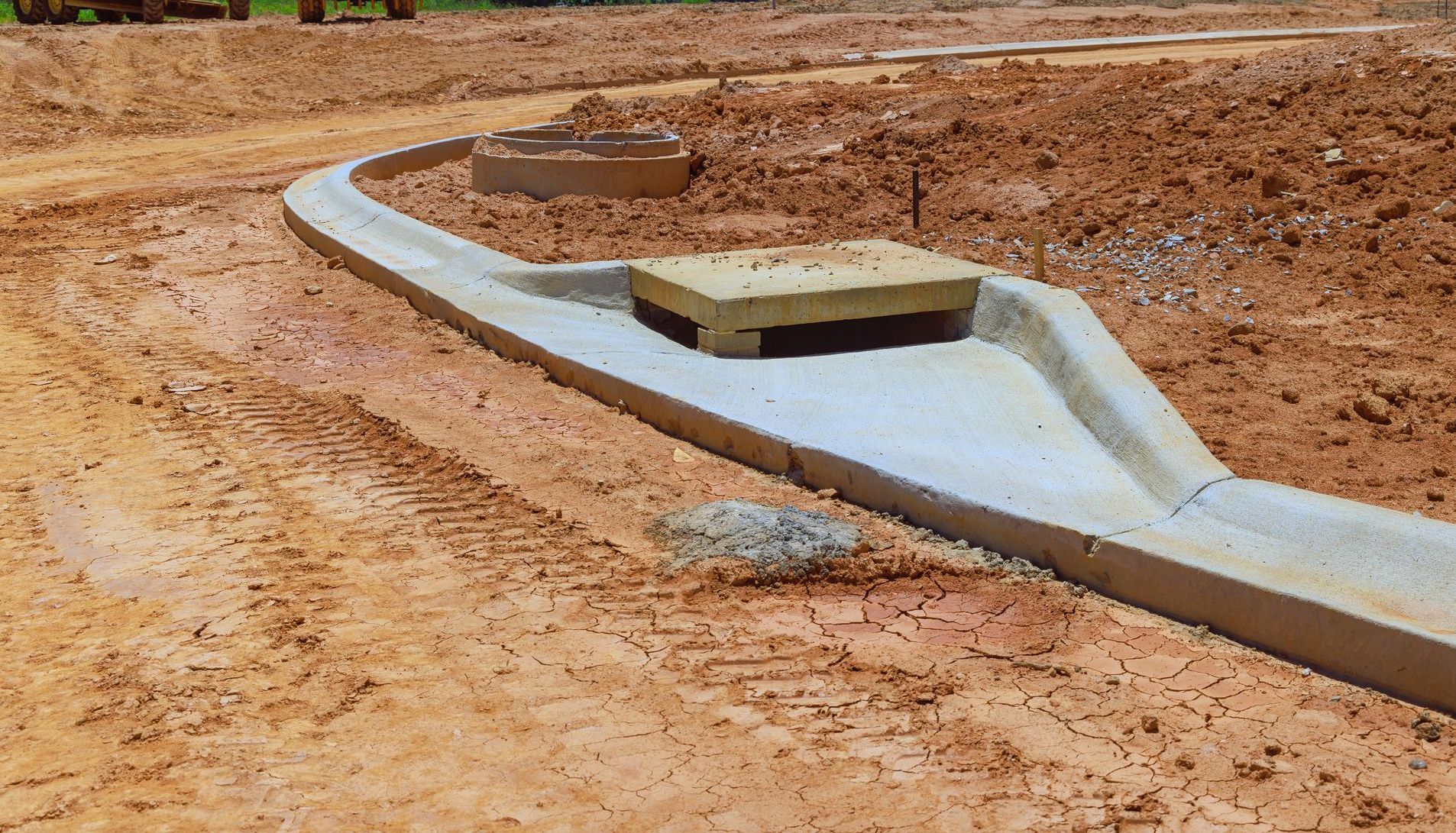 Concrete drainage construction in a dirt setting; curb with a square lid over an opening.