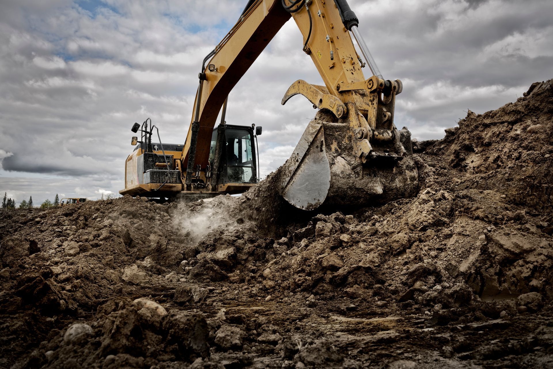 Yellow excavator digging into a pile of brown dirt under a cloudy sky.