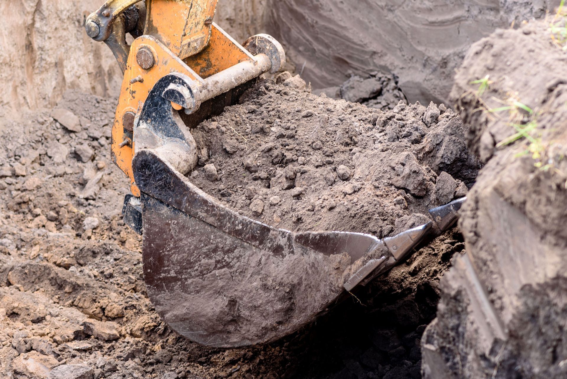 Excavator bucket scooping soil from a trench; construction site with earth tones.