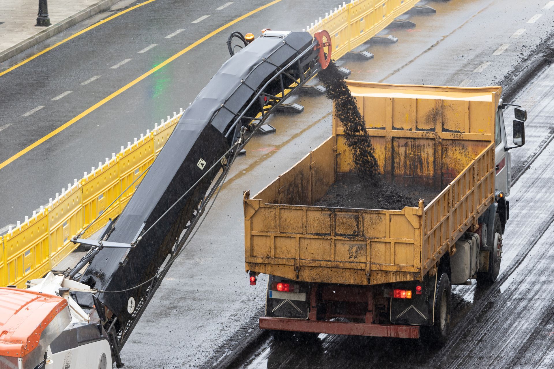 Asphalt milling machine depositing asphalt into an orange dump truck on a city street.
