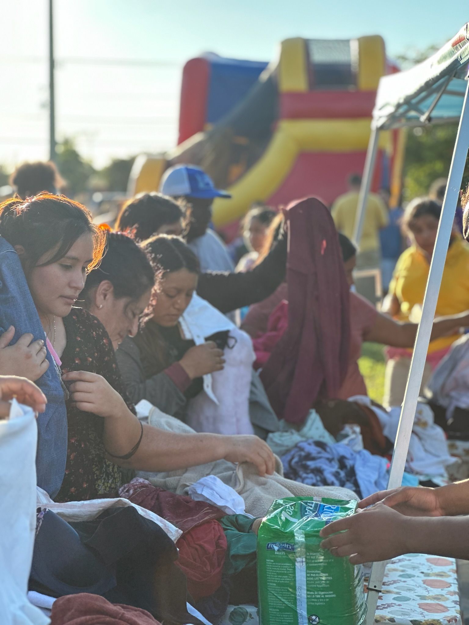 People selecting clothes at an outdoor donation event. Various ethnicities sort through piles of items under a tent; a bounce house is in the background.