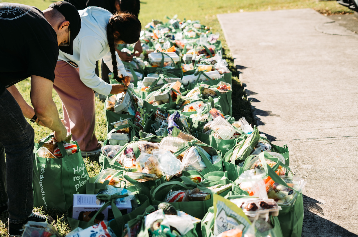 People packing green bags with supplies outdoors on a sunny day. Many filled bags are lined up on the grass.