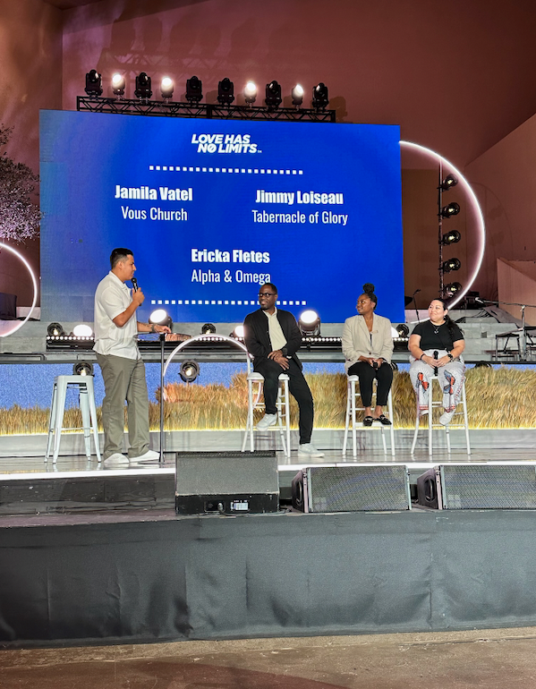Panel discussion on a stage with four people seated, a host standing, and a screen with text,