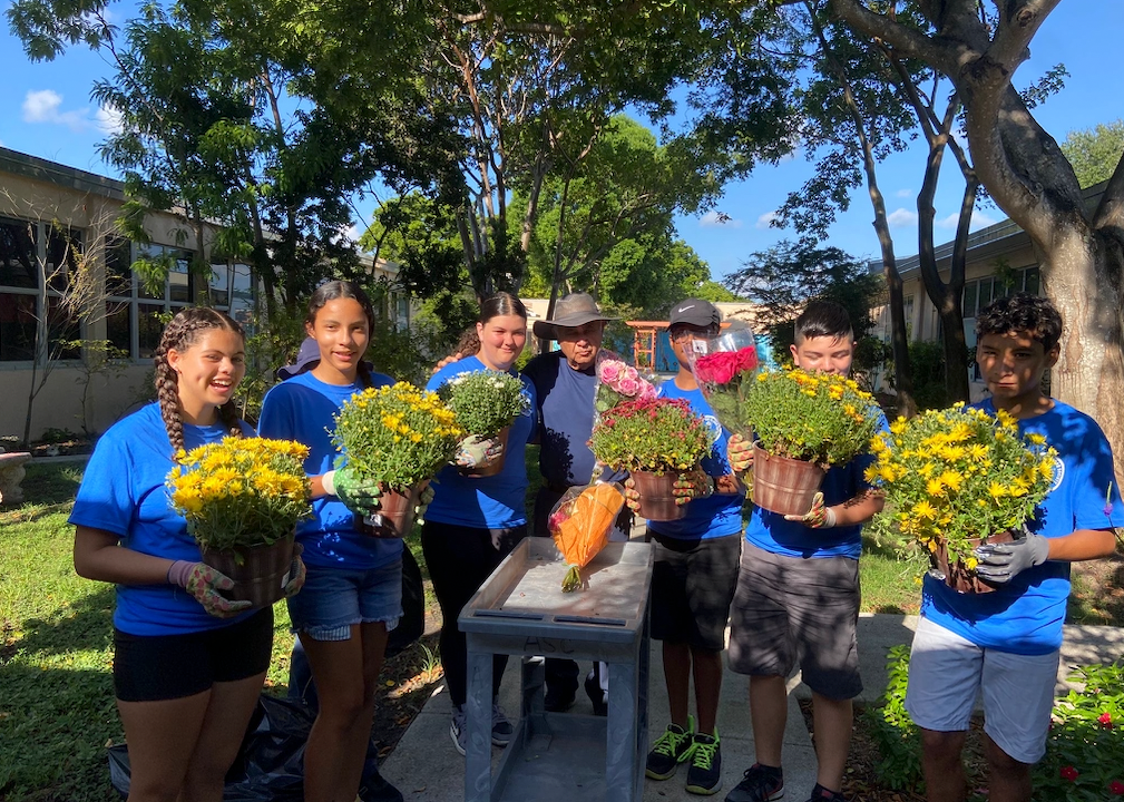 Group of students in blue shirts hold pots of yellow flowers outside. They are gathered near a table.