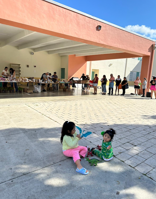 Two young children play on a paved area in front of a building where adults browse tables.