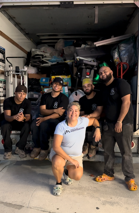 A group of six people smiling in front of a packed moving truck. One person is kneeling, and the others are sitting or standing.
