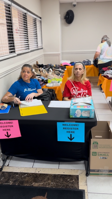 Two women at a registration table, one in blue writing on papers, the other in red. Pink and blue signs say