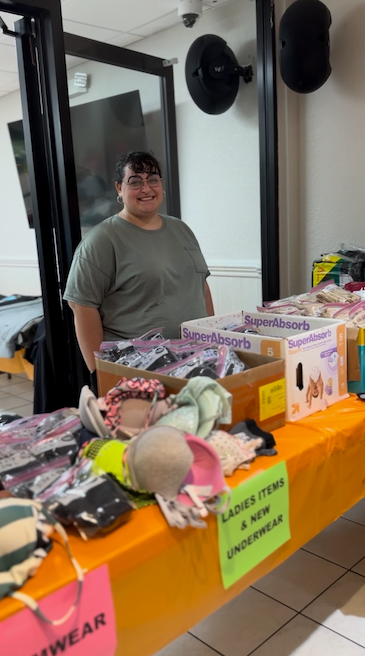 Person smiling behind a table of underwear at an indoor sale. The table is draped in orange, and signs read