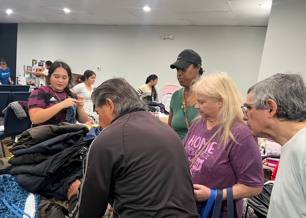People browse clothes at a donation center. Volunteers assist shoppers looking through stacks of items.