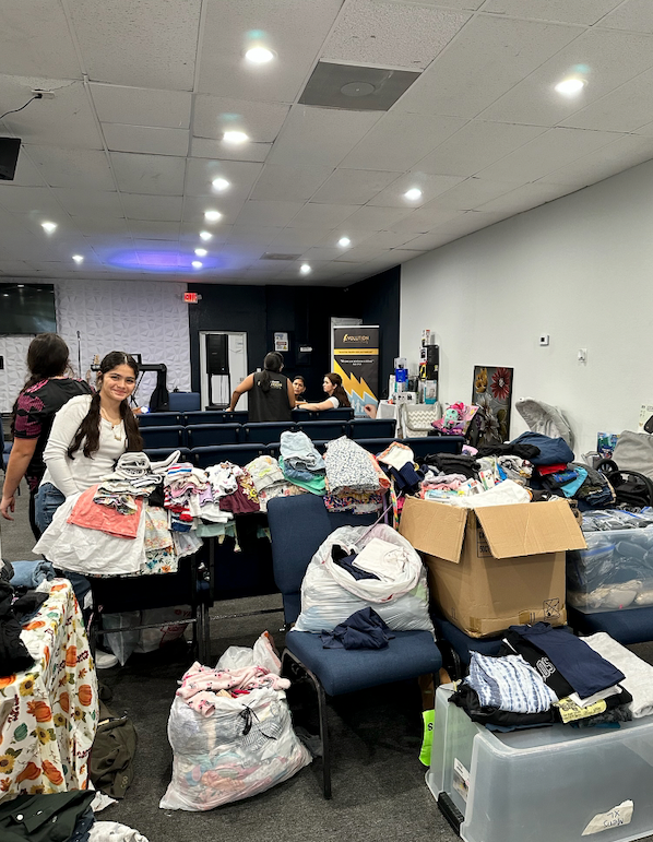 People sorting through clothing in a brightly lit room, possibly a donation or giveaway event. Several piles of clothes and boxes are visible.