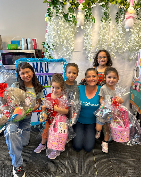 A woman and five children smiling and posing with gift baskets in front of a decorated backdrop. The children are holding Easter-themed baskets and the woman wears a teal shirt.