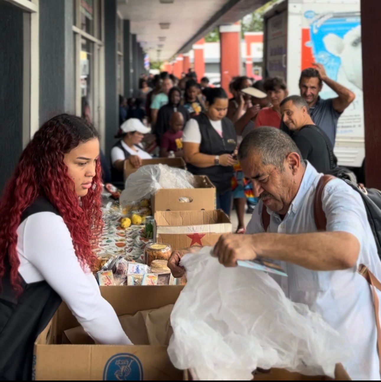 People at a food distribution event. A young woman with red hair and an older man pack food into bags, with a long line of people waiting.