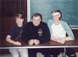Three people seated at a table, in front of a chalkboard. Middle person has fists clenched.