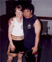 Woman in boxing shorts and man in sleeveless shirt pose together, indoors. Man has arm around woman.