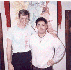 Two men pose for a photo. One with glasses and a light blue shirt. The other wears a white sweatshirt. They stand in front of a colorful painting.