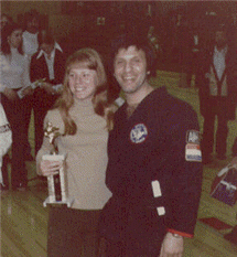 Woman holding trophy with man in a black martial arts uniform, smiling at the camera.