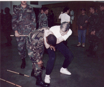 Soldier demonstrating a combat technique. Soldiers in camouflage uniform observe. Indoor setting.