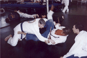 Two people in white gi uniforms grappling on a blue mat, another person observes nearby.