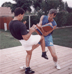Man kicking a pad held by another man outdoors; black shirt, white shorts, blue shorts, sunny day.