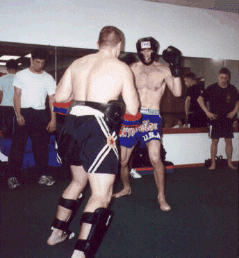Two men in a Muay Thai training session, wearing protective gear, facing each other in a gym.