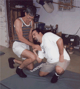 Two men practicing a martial arts hold on a mat. One stands, controlling the other who kneels.