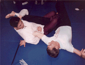 Two people grappling on a blue mat, one in a white shirt applying a choke hold to the other.