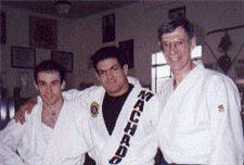 Three men in martial arts uniforms pose together indoors; one in a white gi with