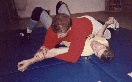 Man in red shirt controlling arm of man in yellow shirt on blue mat.