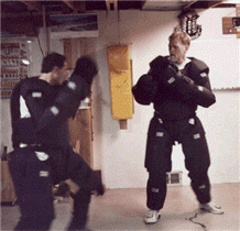 Two people in padded suits sparring in a basement, one throwing a punch, the other blocking.