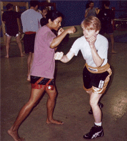 Two people practicing boxing in a gym. One throws a punch, the other blocks. Others watch in the background.