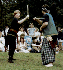 Two people sparring with wooden sticks outdoors, watched by a crowd.