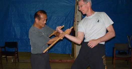 Two men practicing with wooden sticks. One blocks while the other appears to strike in an indoor setting.
