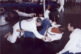Two people in white uniforms on a mat, grappling in a Jiu-Jitsu training session. Others watch.