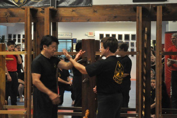 Two people practice Wing Chun hand techniques near a wooden dummy in a martial arts studio.